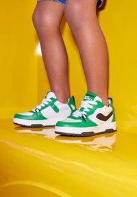 Groene en witte sneakers met zwarte accenten, voorzien van een ronde neus, veters en een gewatteerde kraag; geplaatst tegen een glanzende gele achtergrond.