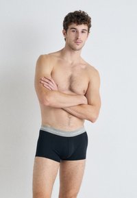 Črne bombažne bokserice zopsko z rjavim pasom Calvin Klein, prilegajoča se zasnova, minimalna raztegljivost in gladka tekstura. Model pozira z rokami prekrižanimi.