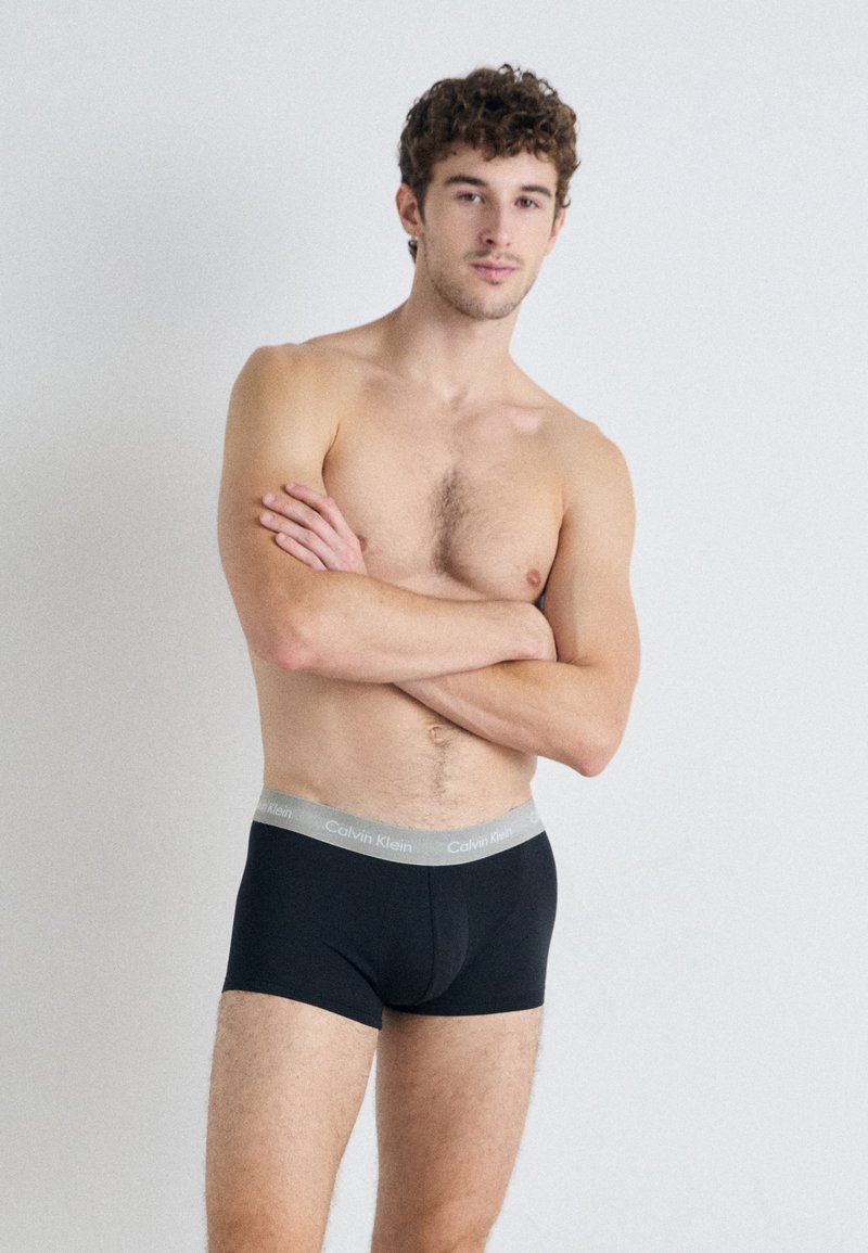 Fekete pamut boxer alsónadrág szürke Calvin Klein logós derékpánttal, testhez simuló kialakítással, minimális rugalmassággal és sima textúrával. A modell karját keresztbe tett állapotban pózol.
