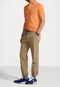 Orange polo shirt, khaki tapered pants, and blue sneakers with white accents. Features a striped belt and textured socks. Casual outfit.