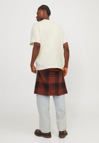 T-shirt beige à manches courtes porté avec une jupe à carreaux noire et rouge sur un jean bleu clair ample et des chaussures marron. Vue de dos.