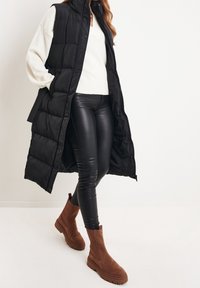 Svart vadderad väst med dragkedja och sidoknappar, bärs över en krämfärgad tröja, kombinerad med svarta läderleggings och bruna mockaankelboots.