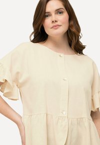 Abito beige chiaro con maniche corte a volant, scollatura rotonda e chiusura con bottoni. Realizzato in tessuto strutturato e presenta una vestibilità ampia.