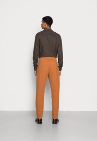 Camisa castanha de manga longa e calças laranja, em mistura de algodão, com um corte descontraído. As calças têm bolsos e bainhas acabadas. Sapatos pretos.
