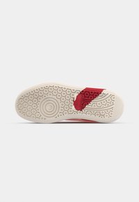 Semelle de chaussure athlétique rouge avec un motif texturé, présentant des grips circulaires et un accent rouge. La semelle est en caoutchouc et a une forme plate.