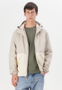 Lichtbeige jas met een capuchon, met een bicolor ontwerp en elastische manchetten. Gedingd over een groene shirt en gecombineerd met blauwe jeans.