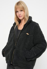 Schwarze Fleecejacke mit einer strukturierten Oberfläche, Reißverschluss und kontrastierendem schwarzen Stoff an den Ärmeln und im Inneren. Mit kleinem Logo.