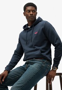 Marine hoodie met een voorzak, trektouwen en een roze logo. Gekozen in combinatie met een spijkerbroek en een gouden horloge op een houten kruk.