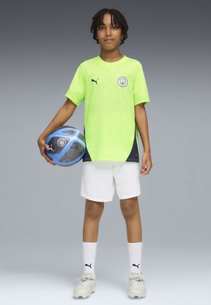 Ung dreng i en neongrøn Manchester City fodboldtrøje, hvide shorts og fodboldstøvler, holder en blå og sort fodbold ved sin side.