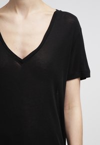 Primer plano de una persona vistiendo una blusa negra de cuello en V semitranslúcida de mangas cortas que muestra el escote y los detalles de la parte superior del pecho.