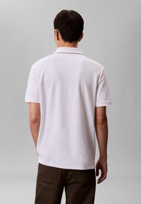 Muž v jednoduchém bílém polokošili s krátkým rukávem a tmavě hnědých kalhotách stojí zády ke kameře na šedém pozadí.