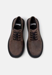 Zapatos de cuero marrón con cordones negros, que cuentan con una suela de goma negra texturizada. En el interior se muestra el logo de la marca "CAMPER".
