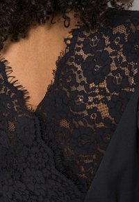 Schwarzes Kleid mit einem Spitzenüberzug mit Blumenmustern und ausgefransten Kanten am Ausschnitt, im Kontrast zu einem glatten Stoff.