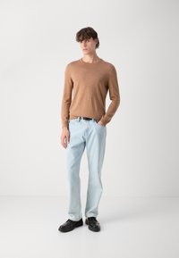 Light brown long-sleeve top, light blue jeans, and black shoes. Simple design, smooth texture. Model stands against a plain white background.