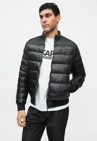 Geacă neagră matlasată cu design orizontal, închidere cu fermoar, manșete și guler rib-knit. Asortată cu un hanorac gri deschis și pantaloni negri.