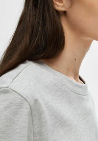 Helles graues Sweatshirt mit Rundhalsausschnitt, strukturiertem Stoff und sichtbaren Nahtdetails. Haare bedecken teilweise den Halsausschnitt.