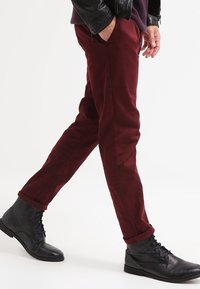 Pantaloni chino color bordeaux in cotone con taglio a gamba dritta, risvolti arrotolati, abbinati a stivaletti neri in pelle con lacci e superficie texturizzata.