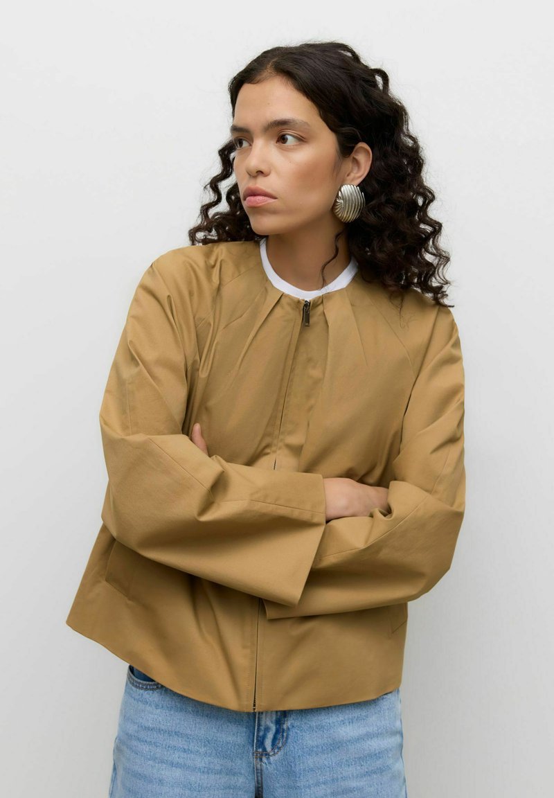 Veste marron courte avec col montant, dotée d'une fermeture éclair frontale et de manches larges. Associée à un jean denim bleu clair. Finition texturée et mate.