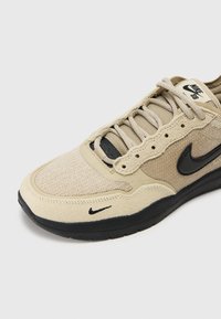 Sapatilhas bege com um upper texturizado que combina malha e camurça. Logotipo preto da Nike, atacadores planos e sola de borracha para aderência.