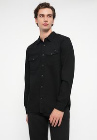 Juoda ilga rankovė marškiniai, pagaminti iš tekstūruoto audinio. Turi dvi krūtinės kišenes ir mygtukų užtraukimą. Prigulanti forma su klasikiniu apykakle.