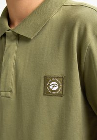 Oliwkowa koszulka polo z teksturowanej tkaniny, z wyszywanym kwadratowym naszywką z logo i nazwą marki w białym i zielonym kolorze.