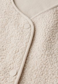 Vêtement beige texturé en tissu pelucheux et bouclé, avec un dessous lisse, bordé d'un trim beige plus doux et fermé par des boutons en plastique.