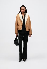 Jachetă de suede de culoare chihlimbar cu căptușeală crem, cu butoane duble, combinată cu un costum negru și o geantă, pe un fundal neutru.