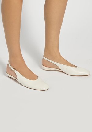 Witte leren slingback platte schoenen met vierkante neuzen en kleine hakken, gedragen op blote voeten tegen een lichtgrijze achtergrond.
