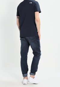 Merinavyinen puuvillat-paita, jossa on logo selässä, yhdistetty tummansinisiin farkkuihin ja valkoisiin tennareihin. Rennon mallinen, jossa on käännetyt hihansuut.