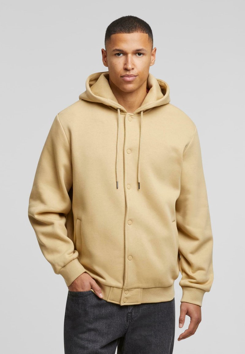 Beige capuchontrui met knoopsluiting, ribgebreide manchetten en zoom, voorzien van trekkoorden en zijzakken. Gladde stoftextuur.