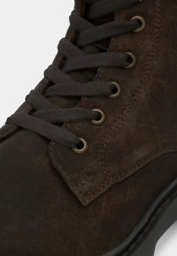 Brūna ādas potīšu zābaki ar tumši pelēkām šņorēm, apaļām metāla acīm un teksturētu virsmu. Aprīkots ar šūtu sānu paneli un gumijas zolīti.