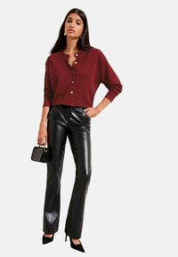 Pulover scurt cu nasturi, texturat și de culoare burgund, asortat cu pantaloni evazați din piele ecologică neagră, cu talie înaltă, și pantofi cu toc cui negri. Geantă mică neagră.