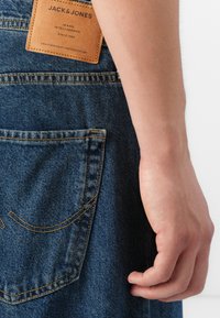 Primer plano de una mano descansando al lado de unos vaqueros de mezclilla azul con una etiqueta de cuero marrón de la marca Jack & Jones en la cintura.