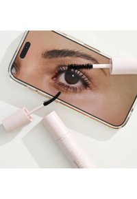 Mascara med dubbla borstar bredvid en telefon som visar en närbild av ett öga med mascara applicerad på övre och nedre fransar.