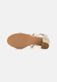 Metalliseeritud kuldne kõrge kontsaga sandaal, ümarate varvastega, ankle strap ja tekstureeritud kummist tallaga. Konts on plokikujuline, tagades stabiilsuse.