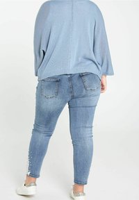 Jasnoniebieski, oversize sweter w połączeniu z dopasowanymi, niebieskimi dżinsami ozdobionymi białą koronką i perłowymi akcentami wzdłuż krawędzi; casualowe trampki.