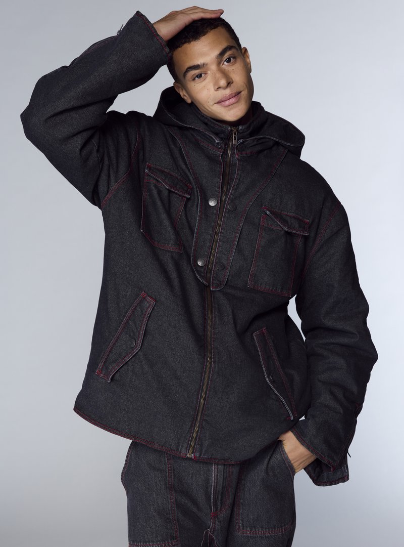 Giacca di denim nera con cuciture rosse, dotata di zip frontale, ampie tasche e cappuccio. Texture liscia con un design a vestibilità comoda.