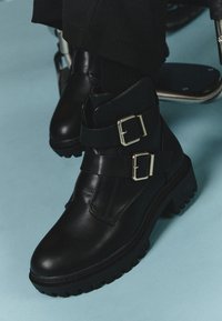 Black leather boots with thick soles and two silver buckles worn with black pants against a light blue floor.