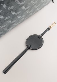 Étiquette de bagage en cuir noir avec étiquette ronde cousue et longue sangle, attachée à la poignée d'une valise grise à motif géométrique.