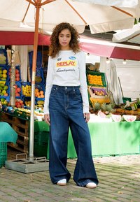En kvinne står under et markedstelt med fargerike frukter og grønnsaker vist bak henne, iført vide jeans og en hvit langermet topp.