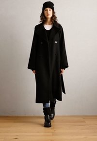 Abrigo de mezcla de lana negro con un diseño de doble botonadura, cuello con muesca y tejido texturizado. Llevado sobre un suéter negro y jeans azules, con botas negras.