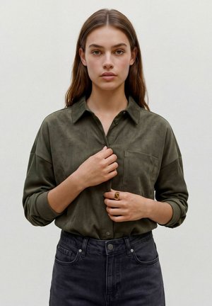Jeune femme aux cheveux bruns portant une chemise en velours côtelé vert olive et un jean foncé, ajustant les boutons de la chemise, fond neutre.