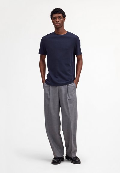 Mand står med hænderne i lommerne iført navyblå kortærmet skjorte, grå vidde bukser og sorte lædersko på hvid baggrund.