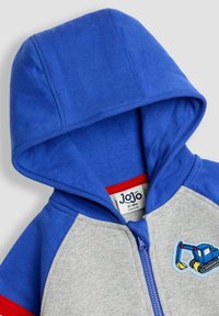 Blauer und grauer Kapuzenpullover mit Reißverschluss, ausgestattet mit einer Fronttasche und einem Stickdetail eines Traktors. Hergestellt aus weichem, strukturiertem Baumwollstoff.