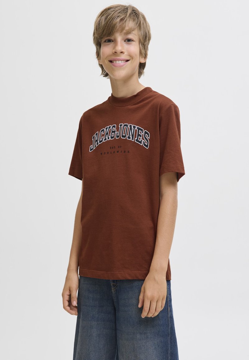 Uśmiechnięty chłopiec ubrany w brązową koszulkę Jack & Jones i niebieskie dżinsowe szorty stoi na tle jednolitego jasnego tła.