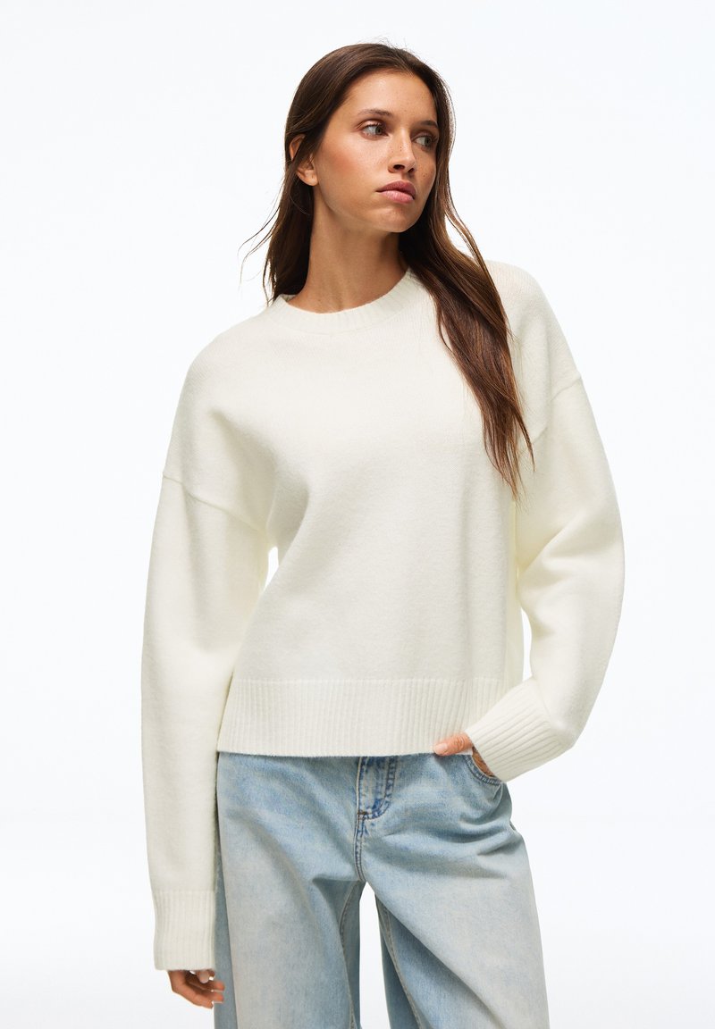 Femme aux longs cheveux bruns portant un pull blanc et un jean bleu clair, debout avec une main dans la poche, regardant sur le côté.