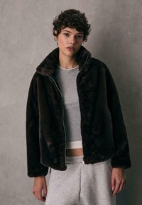 Brun kunstlæderjakke med høj krave, lynlåslukning og blød texture, båret over en grå skjorte og lysegrå sweatpants.
