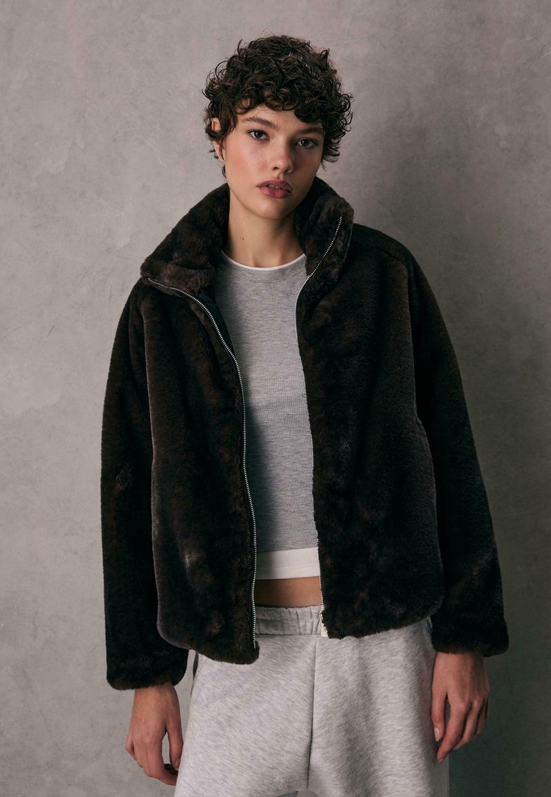 Brun kunstlæderjakke med høj krave, lynlåslukning og blød texture, båret over en grå skjorte og lysegrå sweatpants.