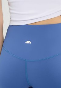 Blaue Leggings mit hoher Taille, die ein dezentes Logo, eine glatte Textur und kontrastierende hellblaue Nähte entlang der Naht und Taille aufweisen.