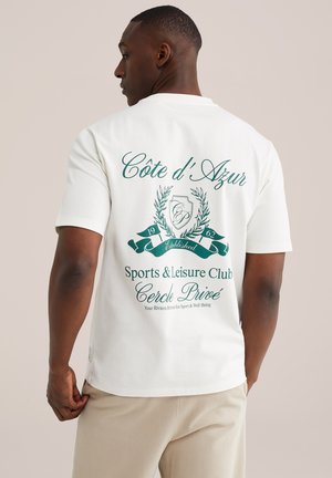 Mężczyzna w białej koszulce z zielonym napisem "Côte d'Azur Sports & Leisure Club Cercle Privé" oraz grafiką na plecach, beżowe spodnie, neutralne tło.
